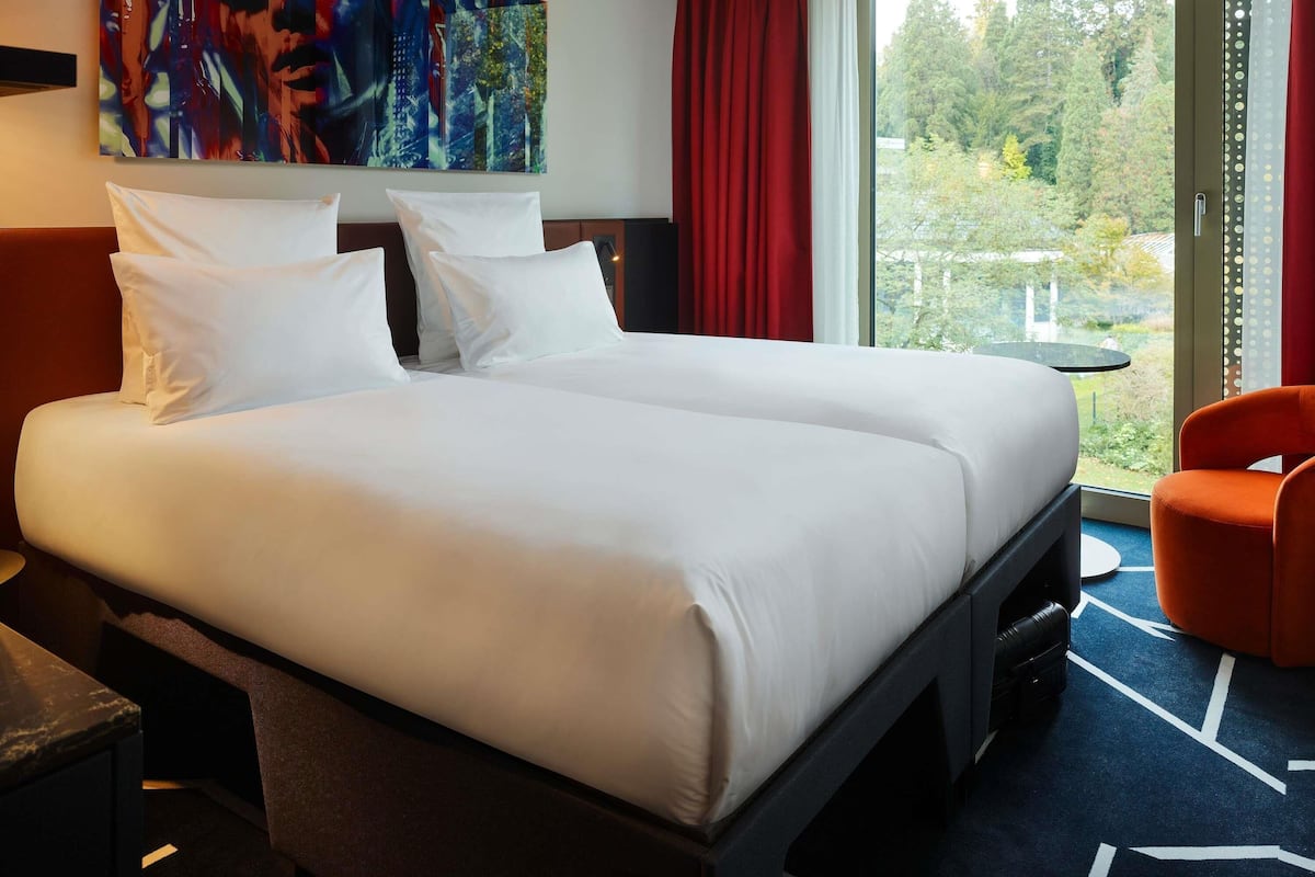 A hotel room with a large bed, a desk, a chair, a painting, and a view of the outdoors.