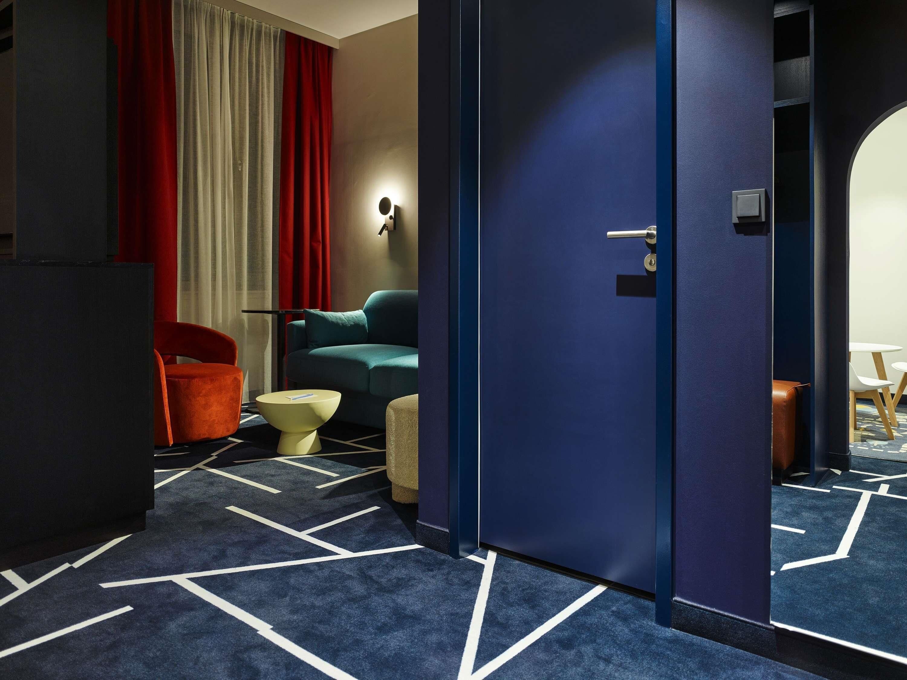 Ein Hotelzimmer mit blauem Teppich, einem türkisfarbenen Sofa, einem roten Sessel, einem kleinen runden Tisch und einer Tür mit Griff.