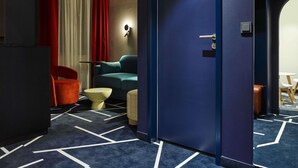 A hotel room with a dark blue carpet, a teal sofa, a red chair, a small round table, and a door with a handle. - TRIBE Baden-Baden (Baden-Baden)