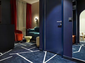 A hotel room with a dark blue carpet, a teal sofa, a red chair, a small round table, and a door with a handle.