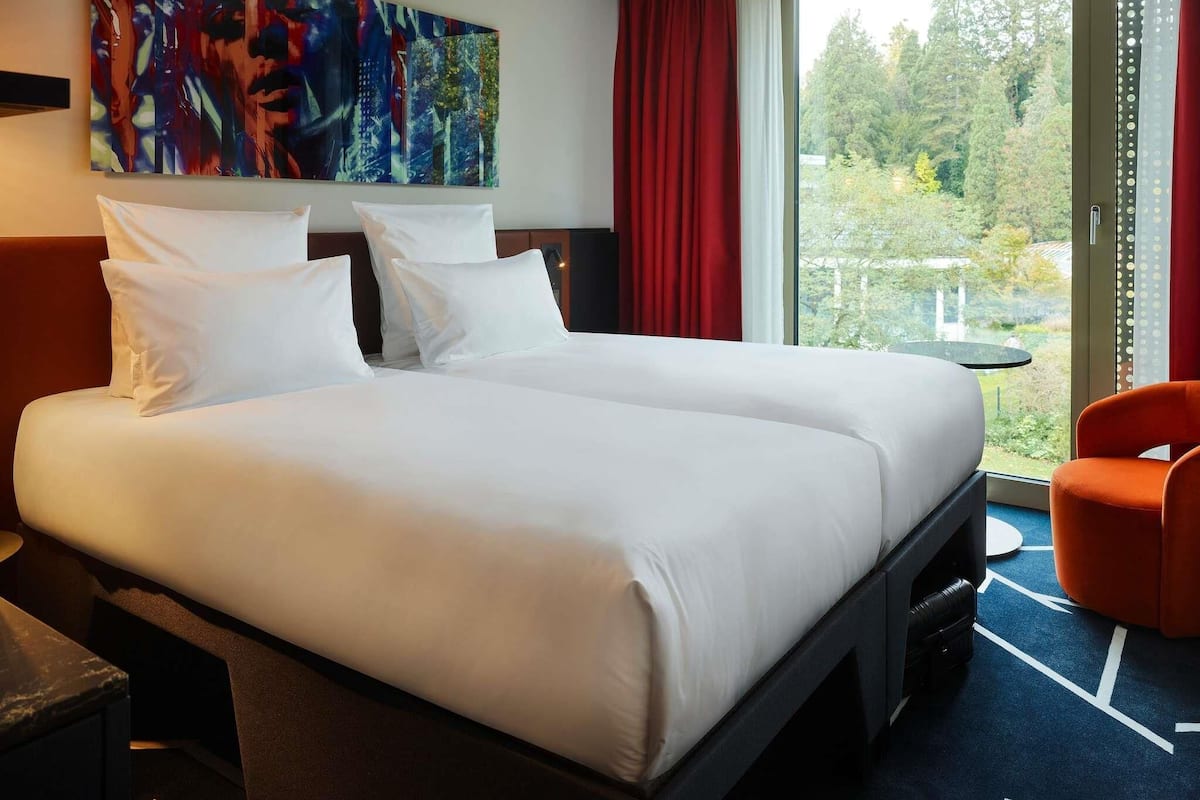 A hotel room with a large bed, a desk, a chair, a painting on the wall, and a view of the outdoors.
