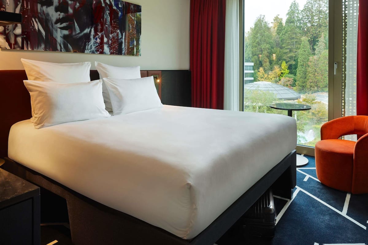 Ein Hotelzimmer mit einem großen Bett, einem Schreibtisch, einem Stuhl, einem Bild an der Wand und Blick auf Bäume durch das Fenster.