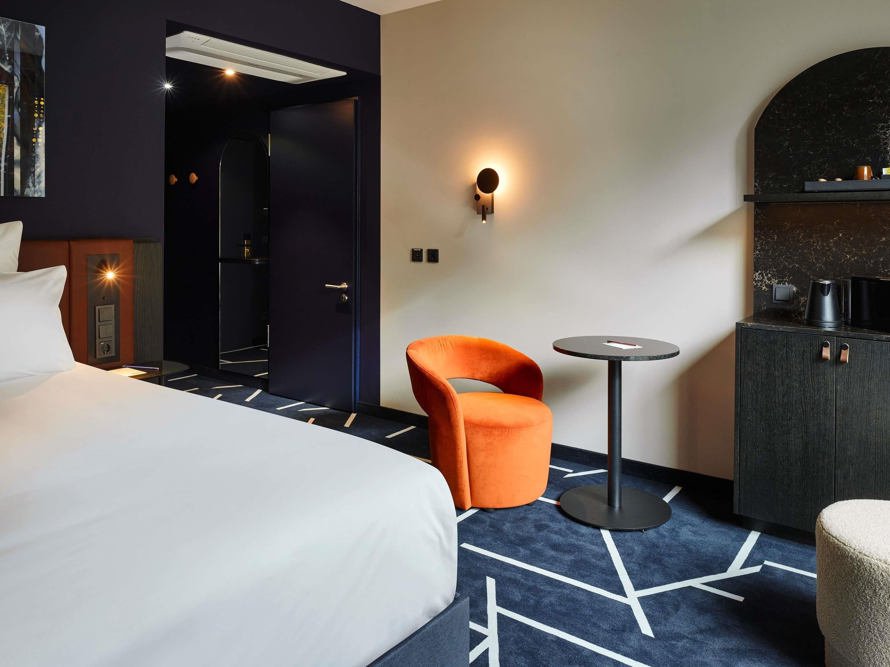 Una habitación de hotel con una cama, una silla naranja, una mesita redonda pequeña, un minibar y una lámpara de pared.
