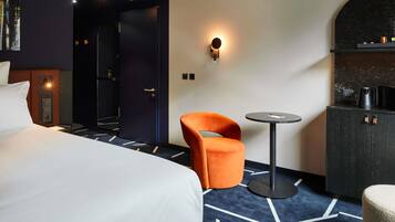Una habitación de hotel con una cama, una silla naranja, una mesita redonda pequeña, un minibar y una lámpara de pared.