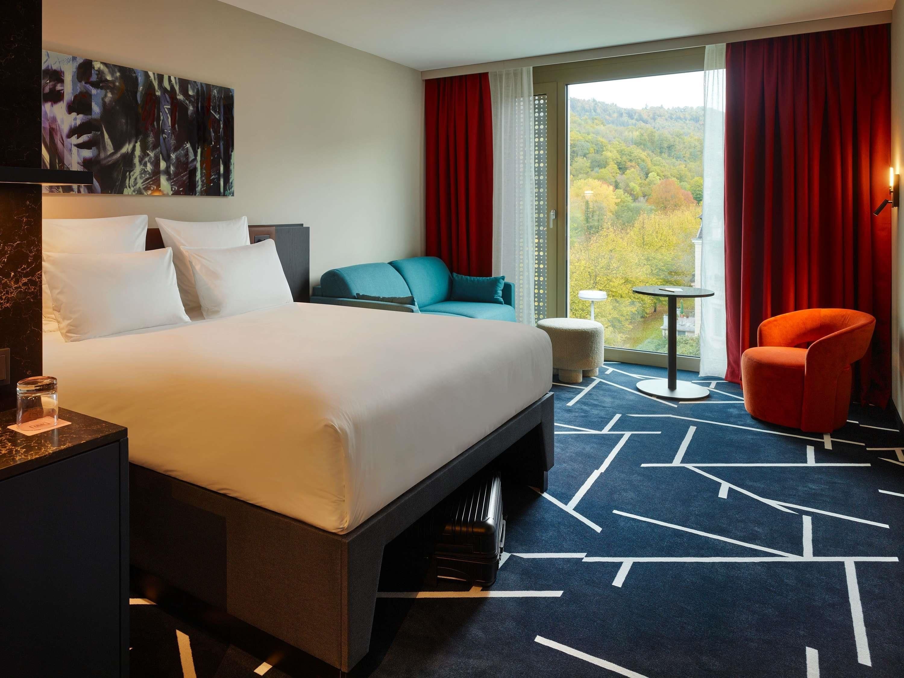 A hotel room with a large bed, a desk with a lamp, a chair, and a view of a forest through a window.