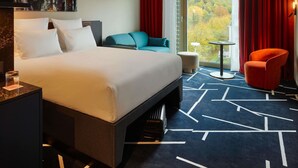 A hotel room with a large bed, a desk with a lamp, a chair, and a view of a forest through a window.