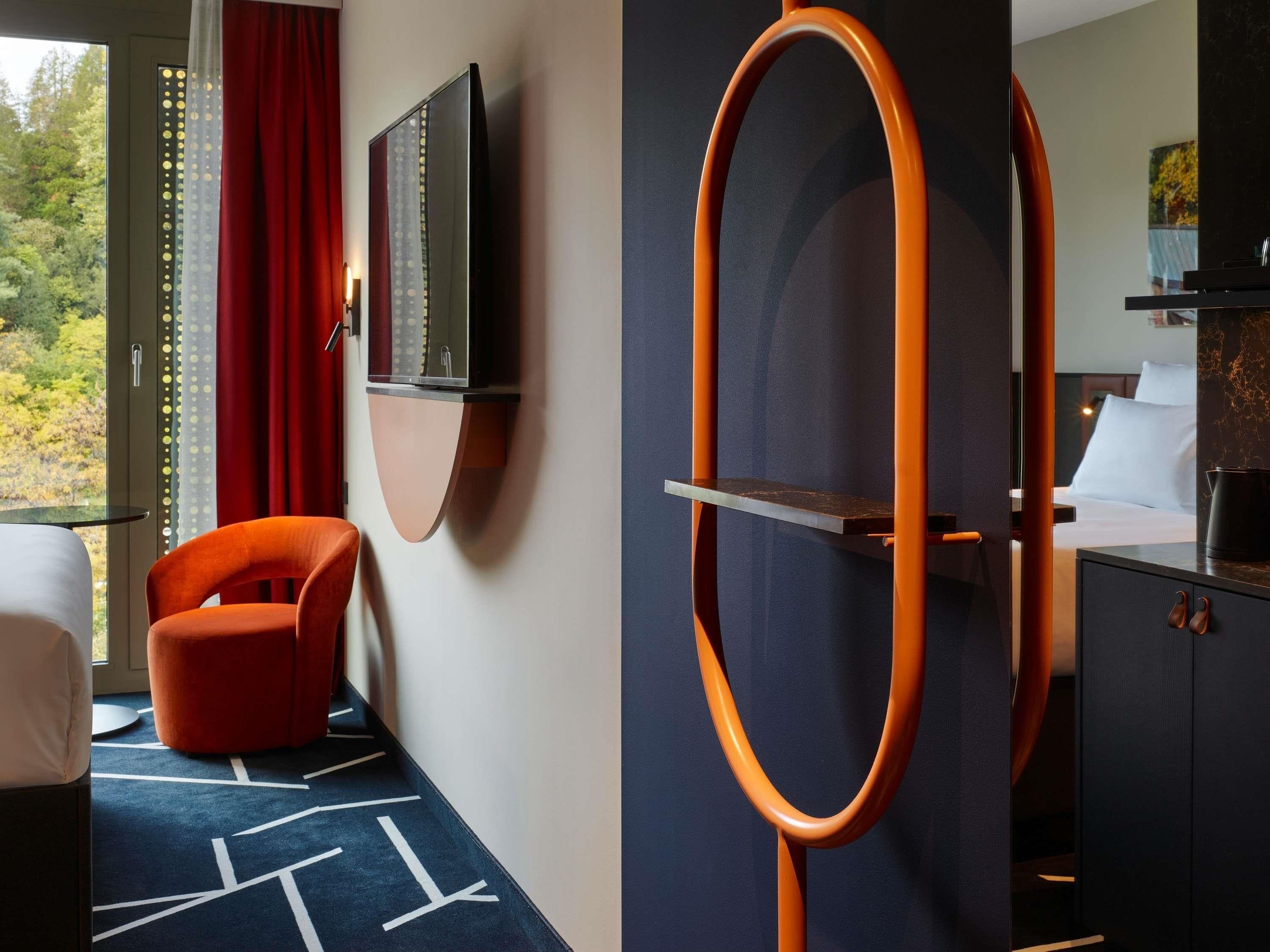 Ein Hotelzimmer mit einem Bett, einem an der Wand befestigten Fernseher, einem roten Sessel und einem großen Spiegel mit orangefarbenem Rahmen.
