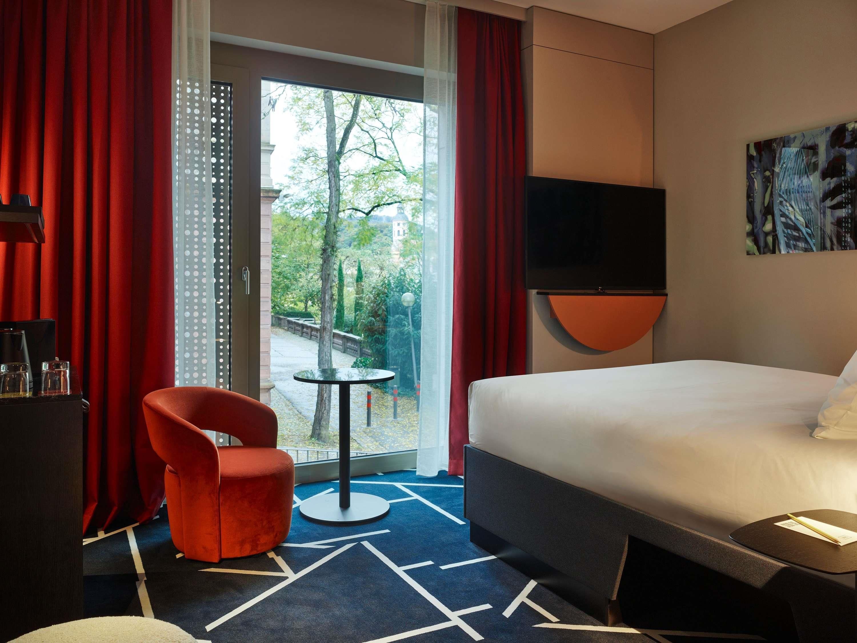 Ein Hotelzimmer mit Bett, einem roten Sessel, einem kleinen Tisch, einem Fernseher und Blick ins Freie.