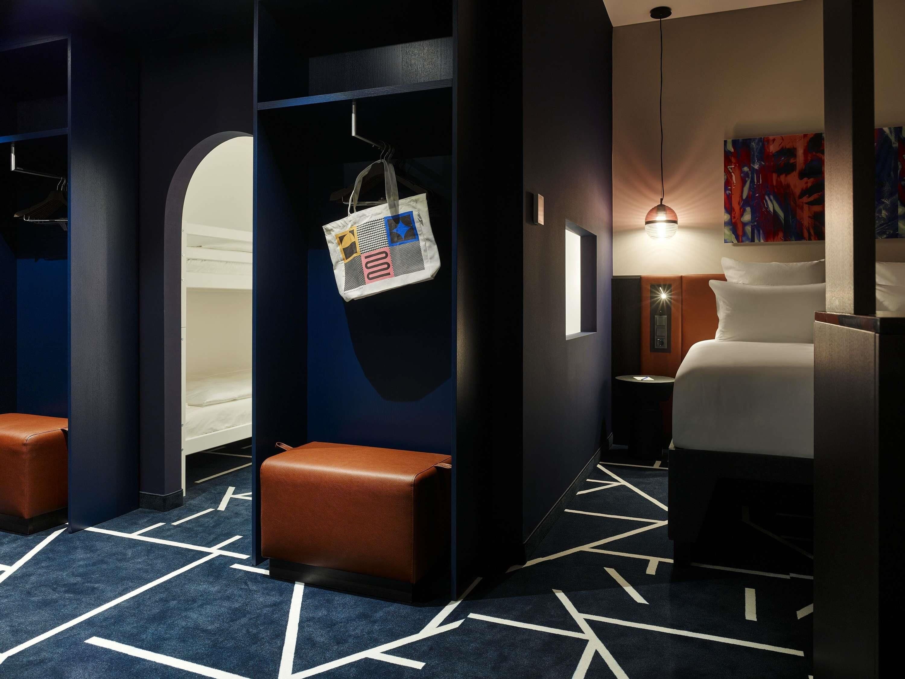 A hotel room with a bed, a desk, a chair, a hanging bag, and a wall-mounted lamp.