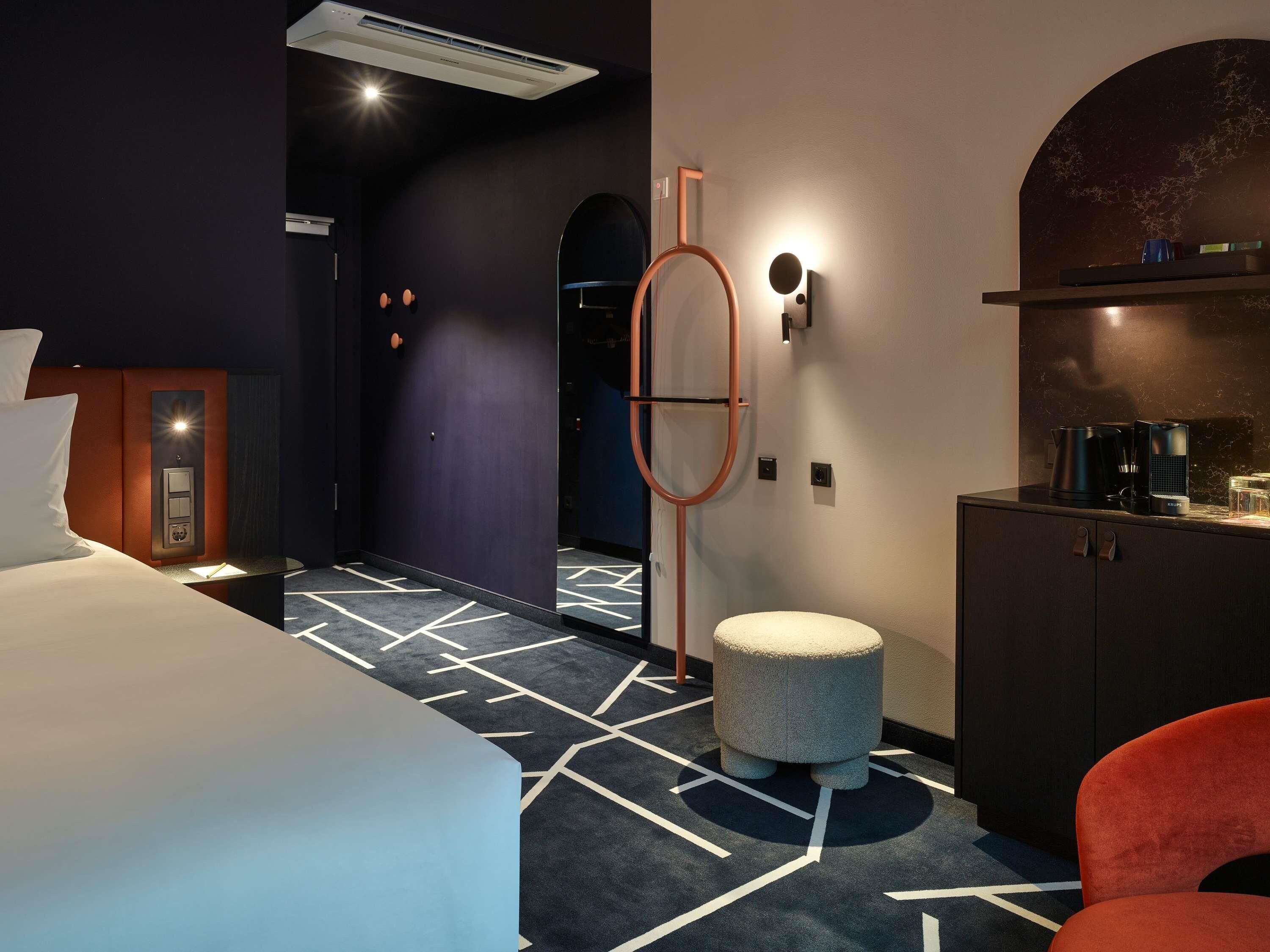 Ein modernes Hotelzimmer mit einem Bett, einem kleinen runden Hocker, einem roten Sessel, einer Kaffeestation und einem wandmontierten Spiegel.