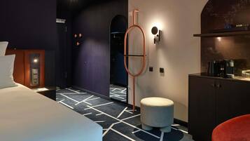 Ein modernes Hotelzimmer mit einem Bett, einem kleinen runden Hocker, einem roten Sessel, einer Kaffeestation und einem wandmontierten Spiegel.