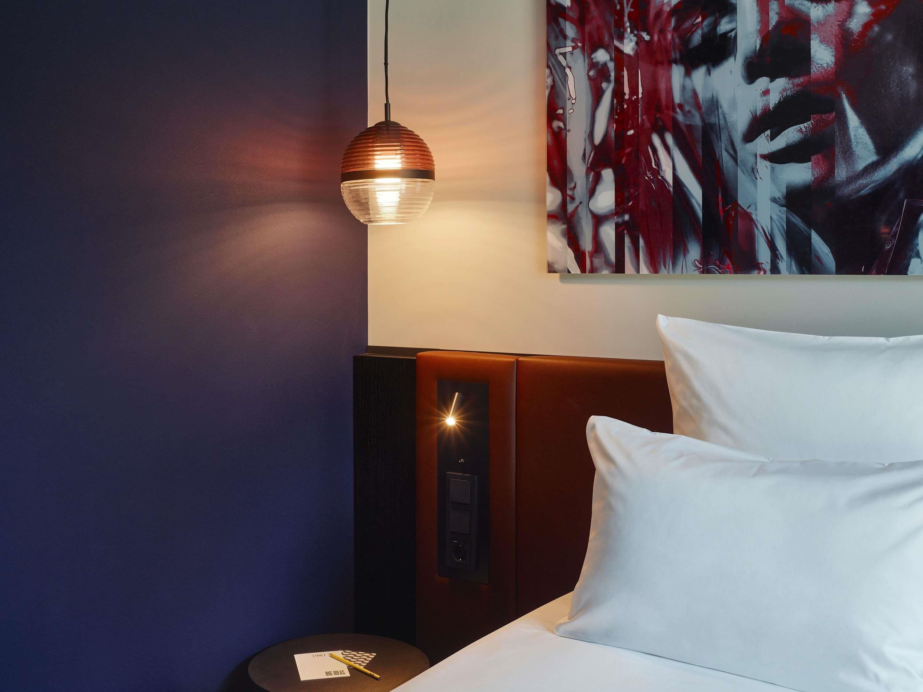 Une chambre d’hôtel avec un lit, une œuvre d’art accrochée au mur, un luminaire suspendu rond et une petite table sur laquelle se trouvent une télécommande et un bloc-notes.