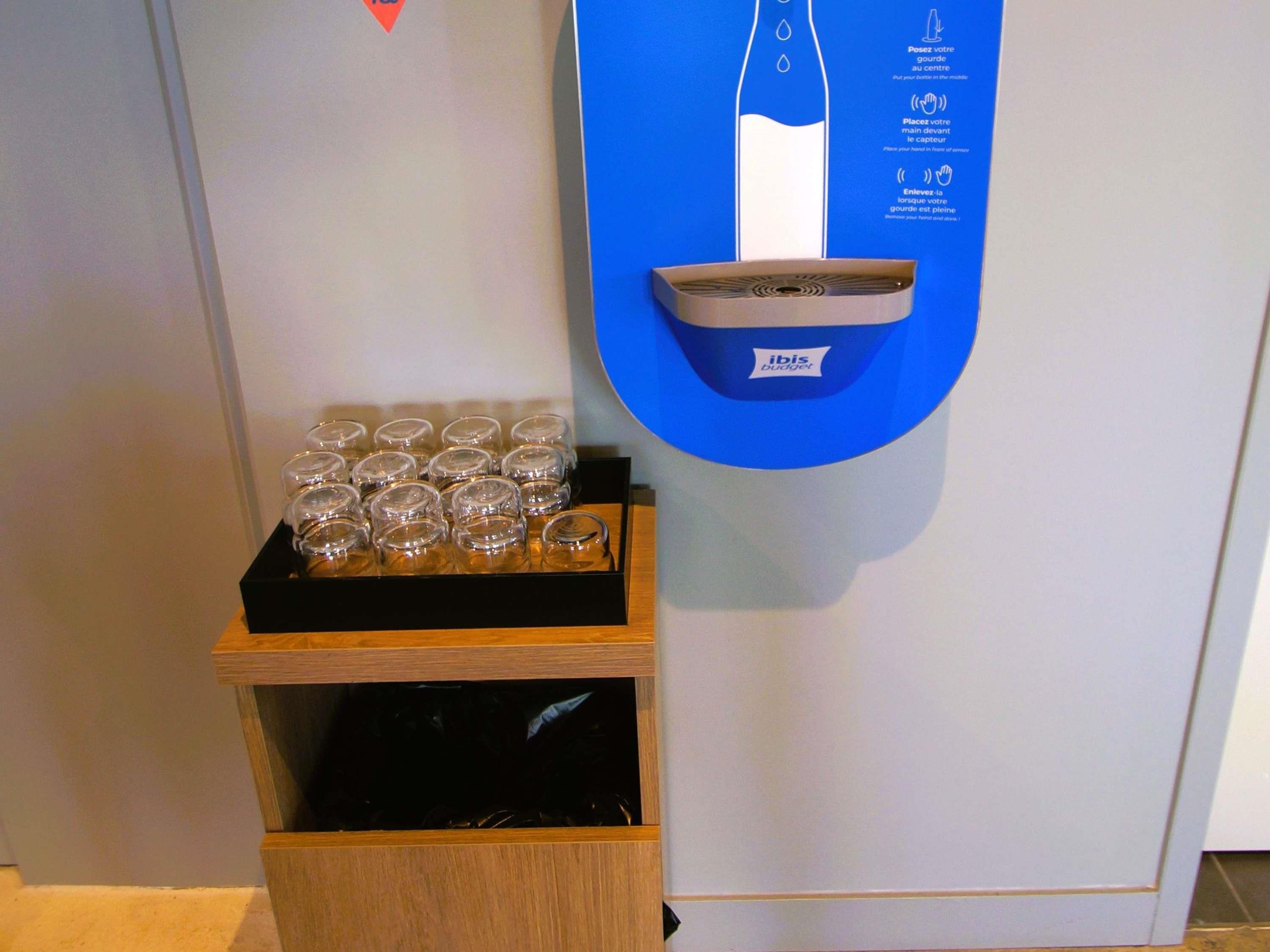 A self-service beverage dispenser with instructions, a tray of glasses, and a wooden storage unit.