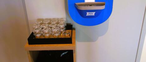 A self-service beverage dispenser with instructions, a tray of glasses, and a wooden storage unit.