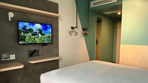 A hotel room with a bed, a mounted TV displaying a nature scene, a small shelf with a pillow, and a door leading to another room.