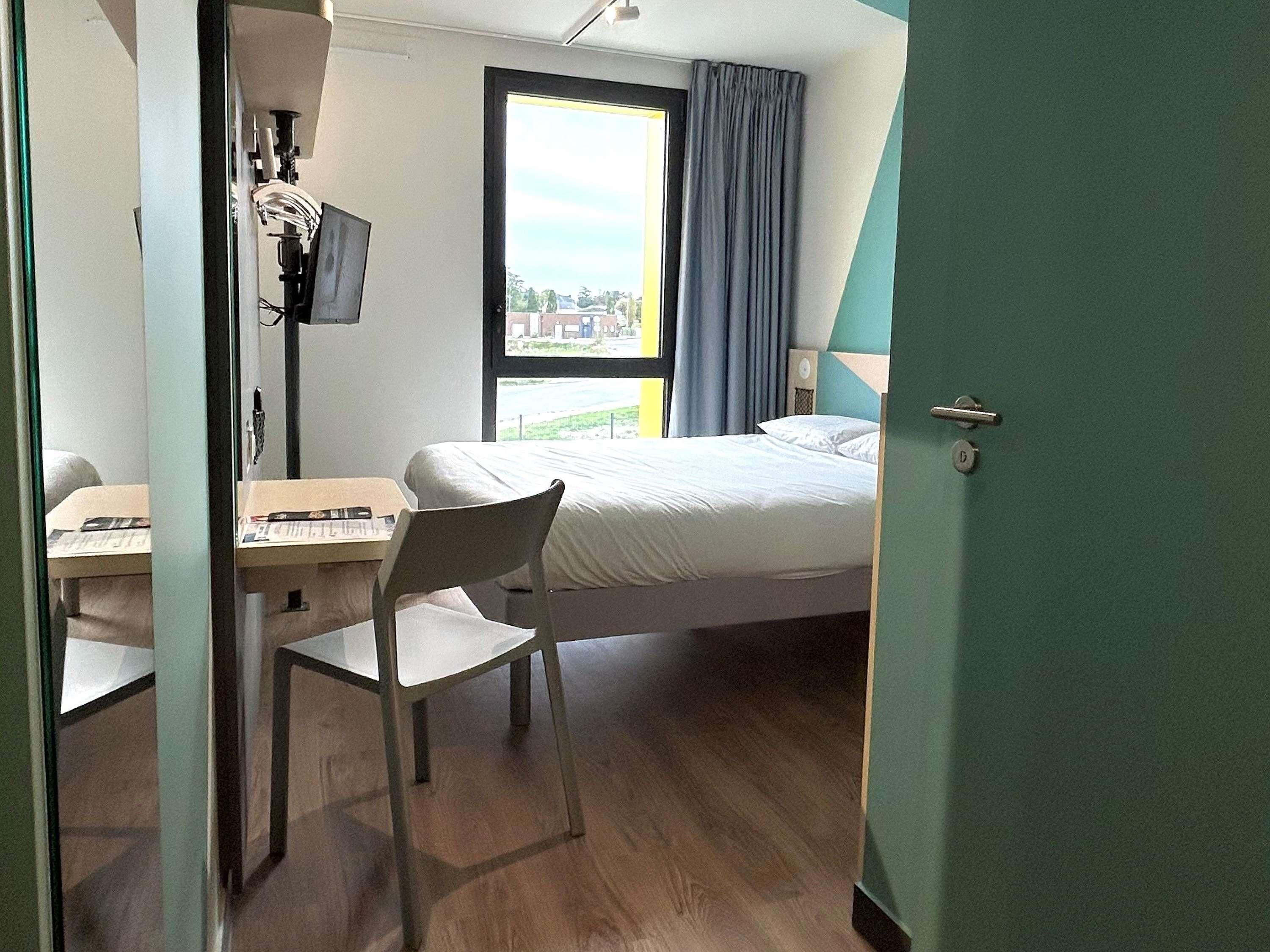Una stanza con un letto, una scrivania e una sedia. Si vede una finestra con tende e una porta.