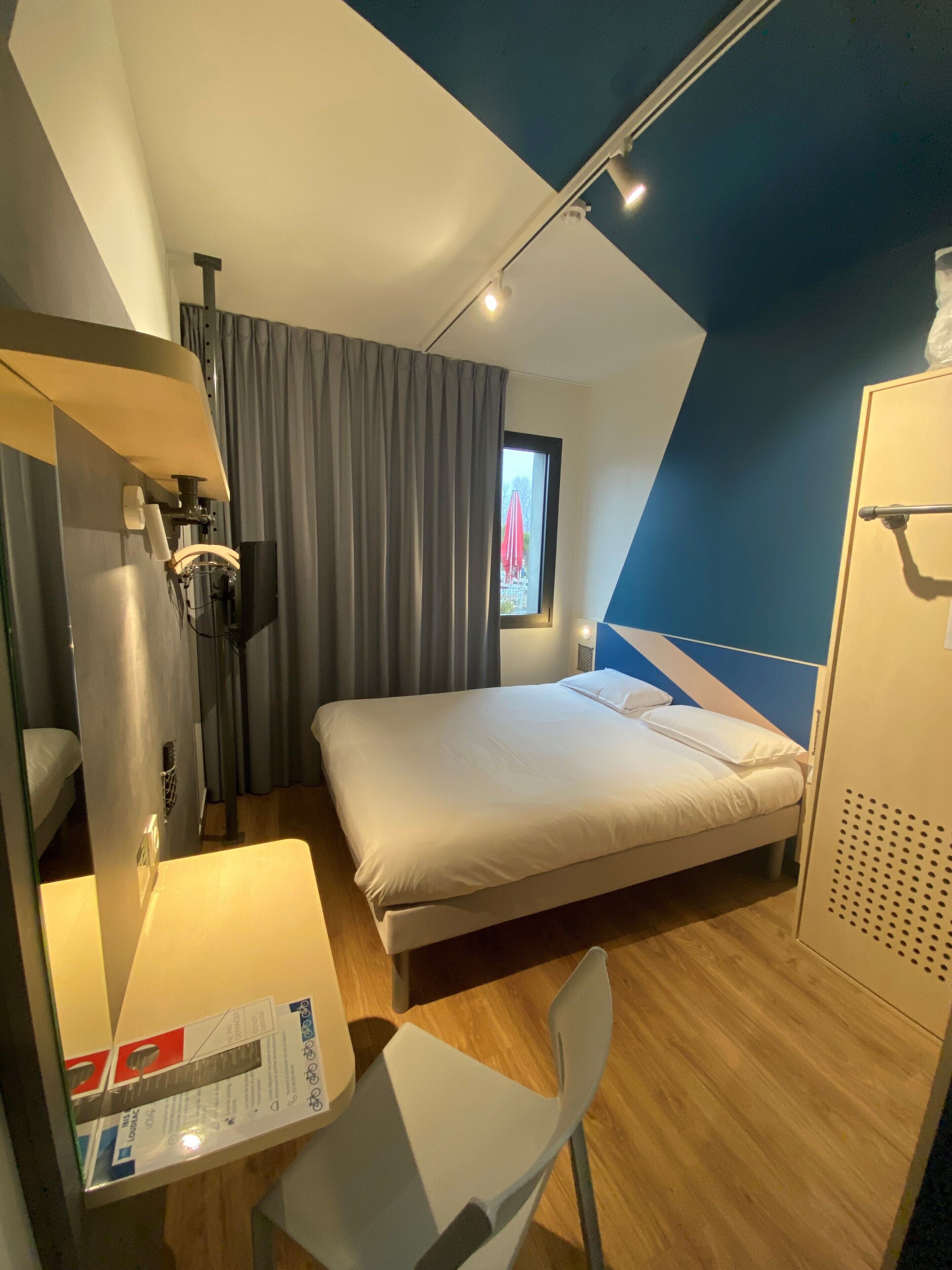 Una habitación de hotel moderna con una cama, un escritorio y una silla. Cuenta con una ventana con cortinas, un televisor y un armario.