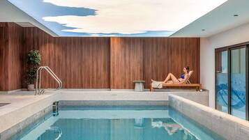 A modern indoor pool with a clear blue water surface, a wooden wall, and a person relaxing on a lounge chair.