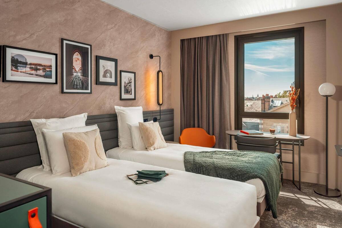 Ein Hotelzimmer mit zwei Betten, einem Schreibtisch, einem Stuhl, einem Fenster mit Blick und gerahmten Bildern an der Wand.