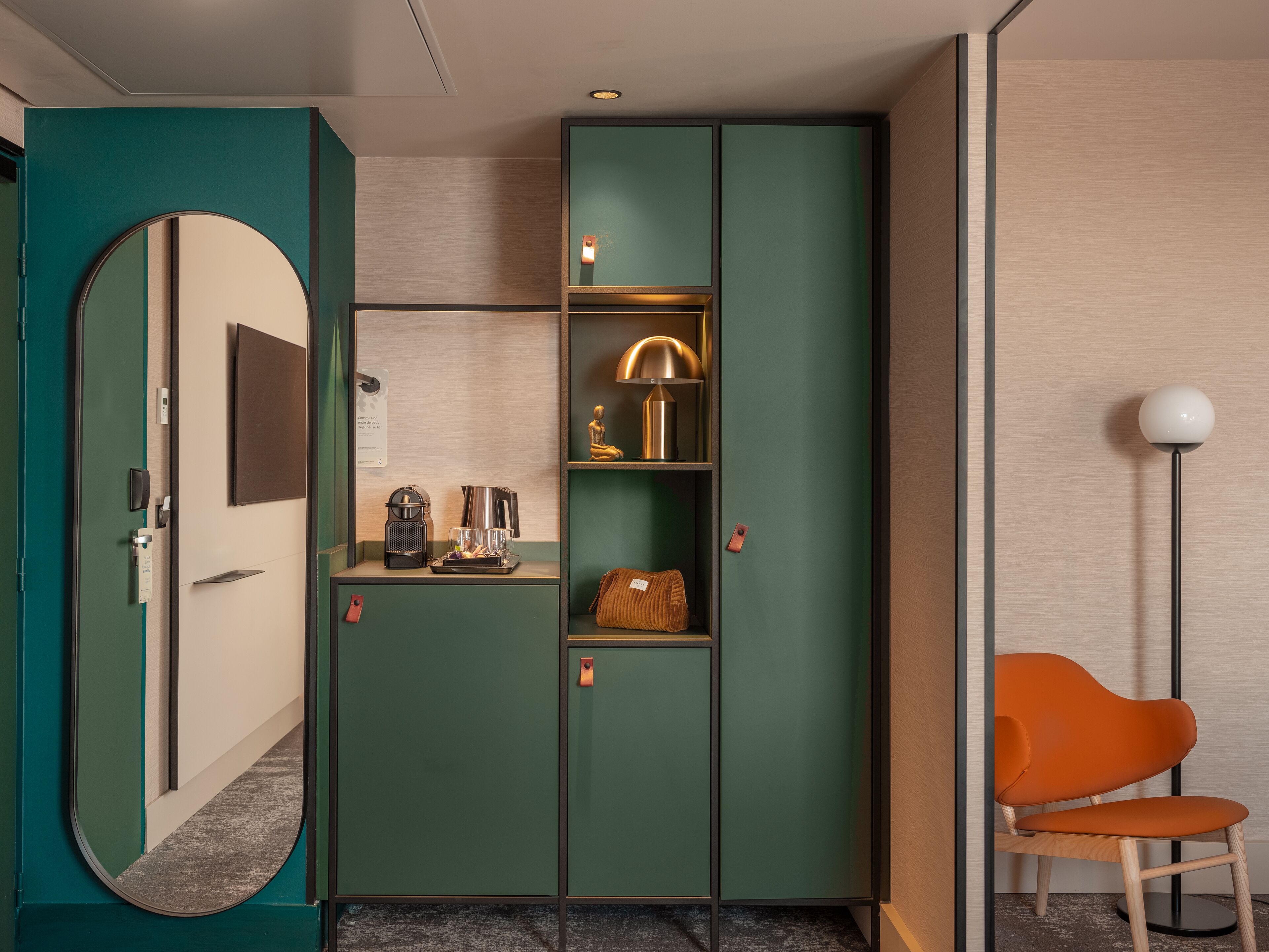 A hotel room with a green wardrobe, a mirror, a coffee machine, a lamp, and an orange chair.