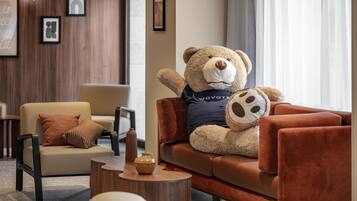 A plush bear on a brown sofa in a modern lobby with a wooden wall, a chair, a small table, and framed pictures.