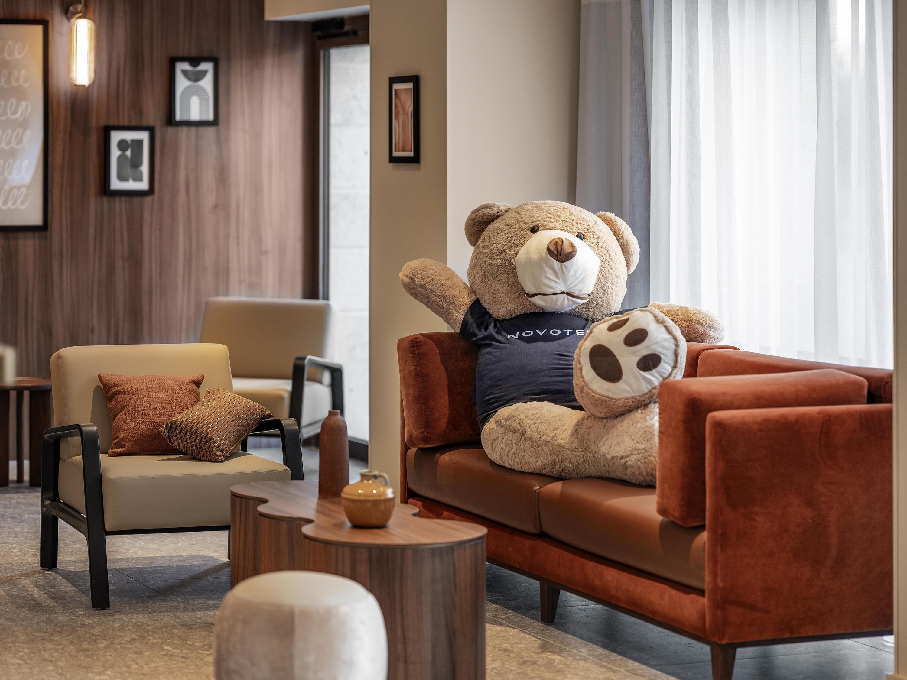 Un oso de peluche sobre un sofá marrón en un vestíbulo moderno con una pared de madera, una silla, una mesita y cuadros enmarcados.