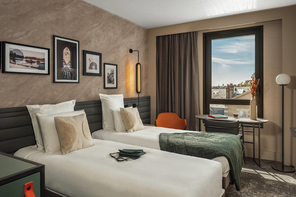 Ein Hotelzimmer mit zwei Betten, einem Schreibtisch, einem Stuhl, einem Fenster mit Aussicht und gerahmten Bildern an der Wand.