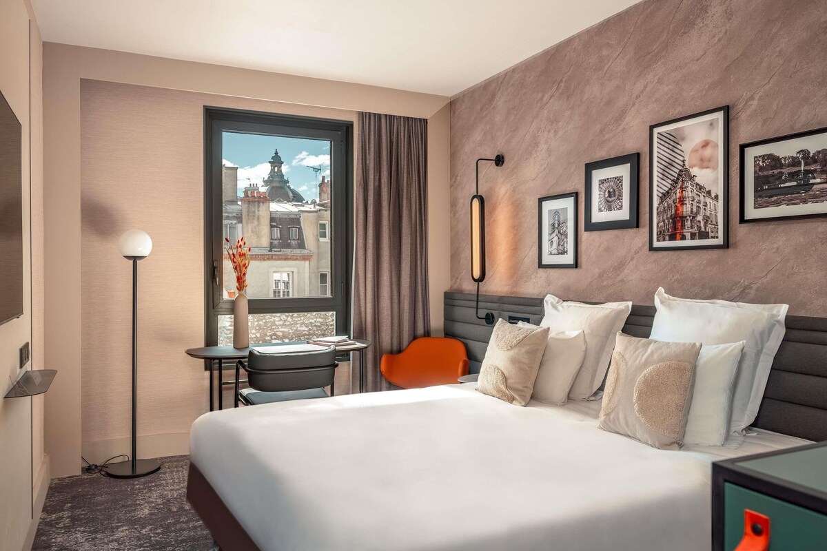 Une chambre d’hôtel avec un grand lit, un bureau avec une chaise, une lampe sur pied, une fenêtre donnant sur des immeubles et des tableaux encadrés accrochés au mur.
