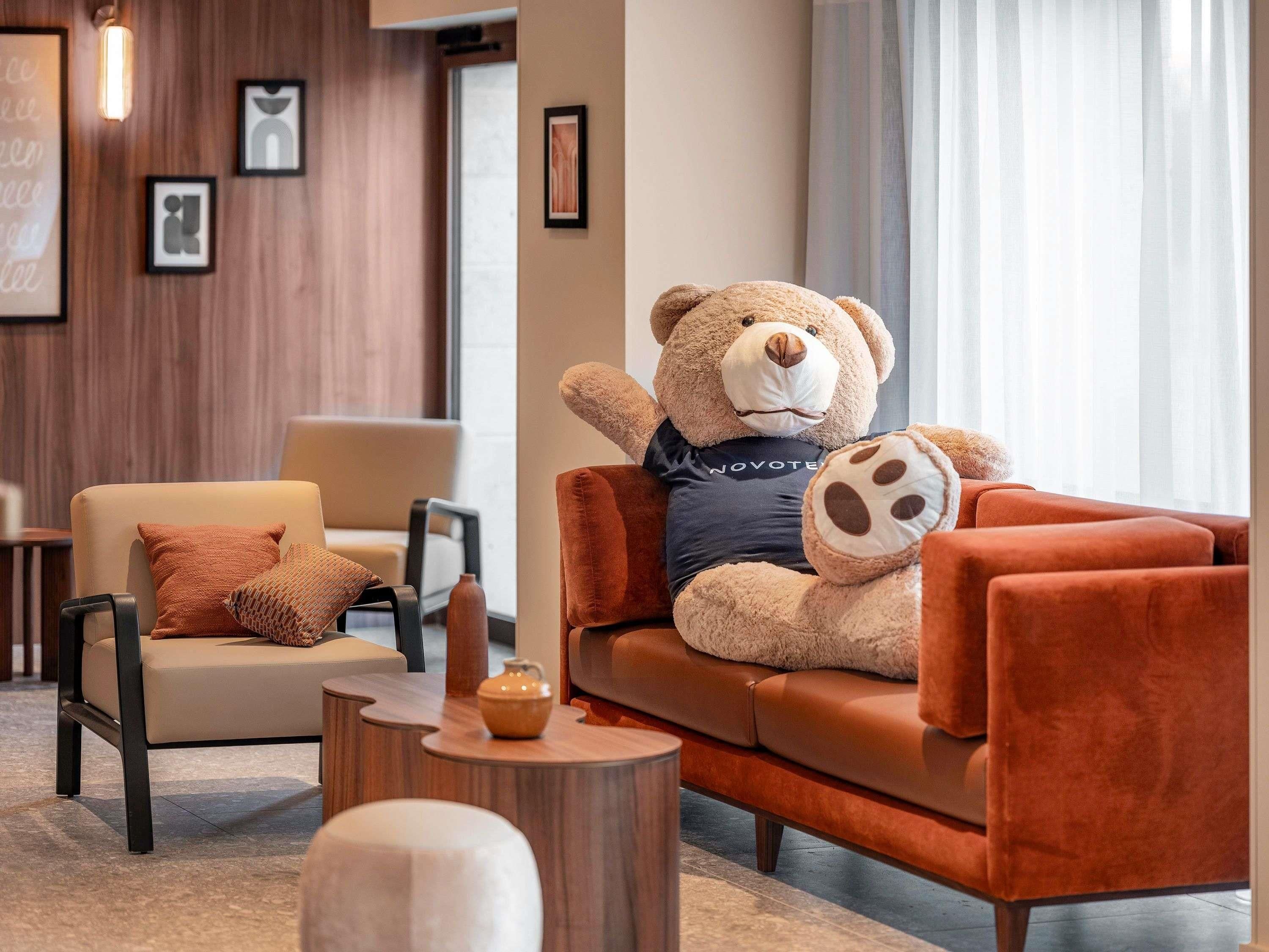 A plush bear sitting on a sofa in a modern lounge area with a wooden wall and framed artwork.