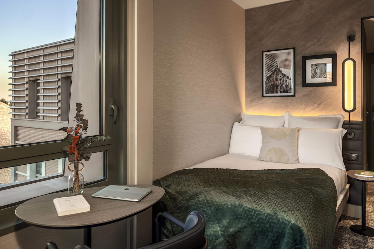 Ein Hotelzimmer mit Bett, Schreibtisch mit Laptop, Blumenvase und Fenster mit Blick auf Gebäude.