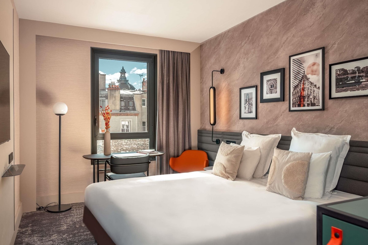 Een hotelkamer met een groot bed, een bureau met stoel, een staande lamp, een raam met uitzicht op gebouwen en lijstwerken aan de muur.