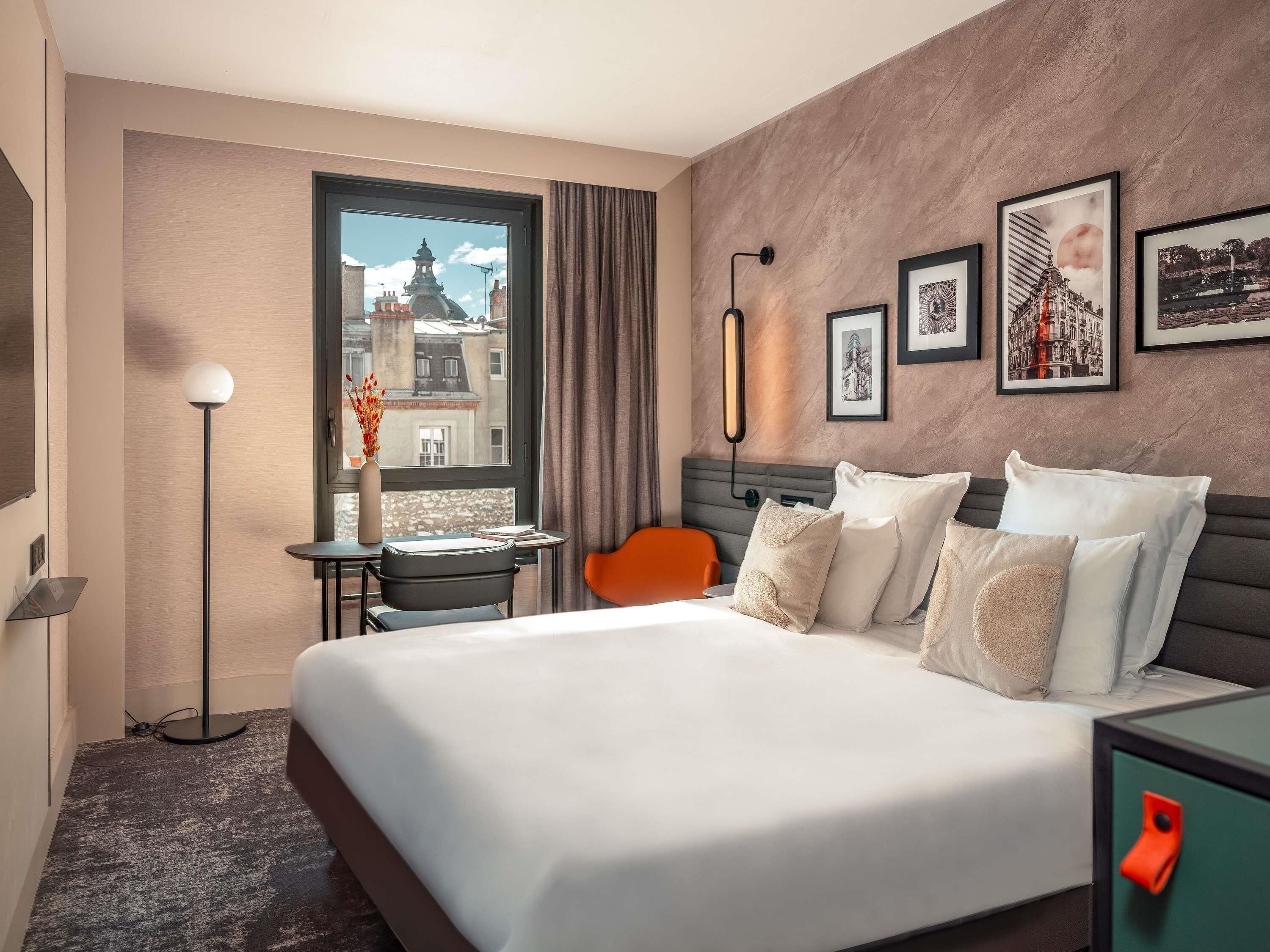 Ein Hotelzimmer mit einem großen Bett, einem Schreibtisch mit Stuhl, einer Stehlampe, einem Fenster mit Blick auf Gebäude und gerahmten Bildern an der Wand.