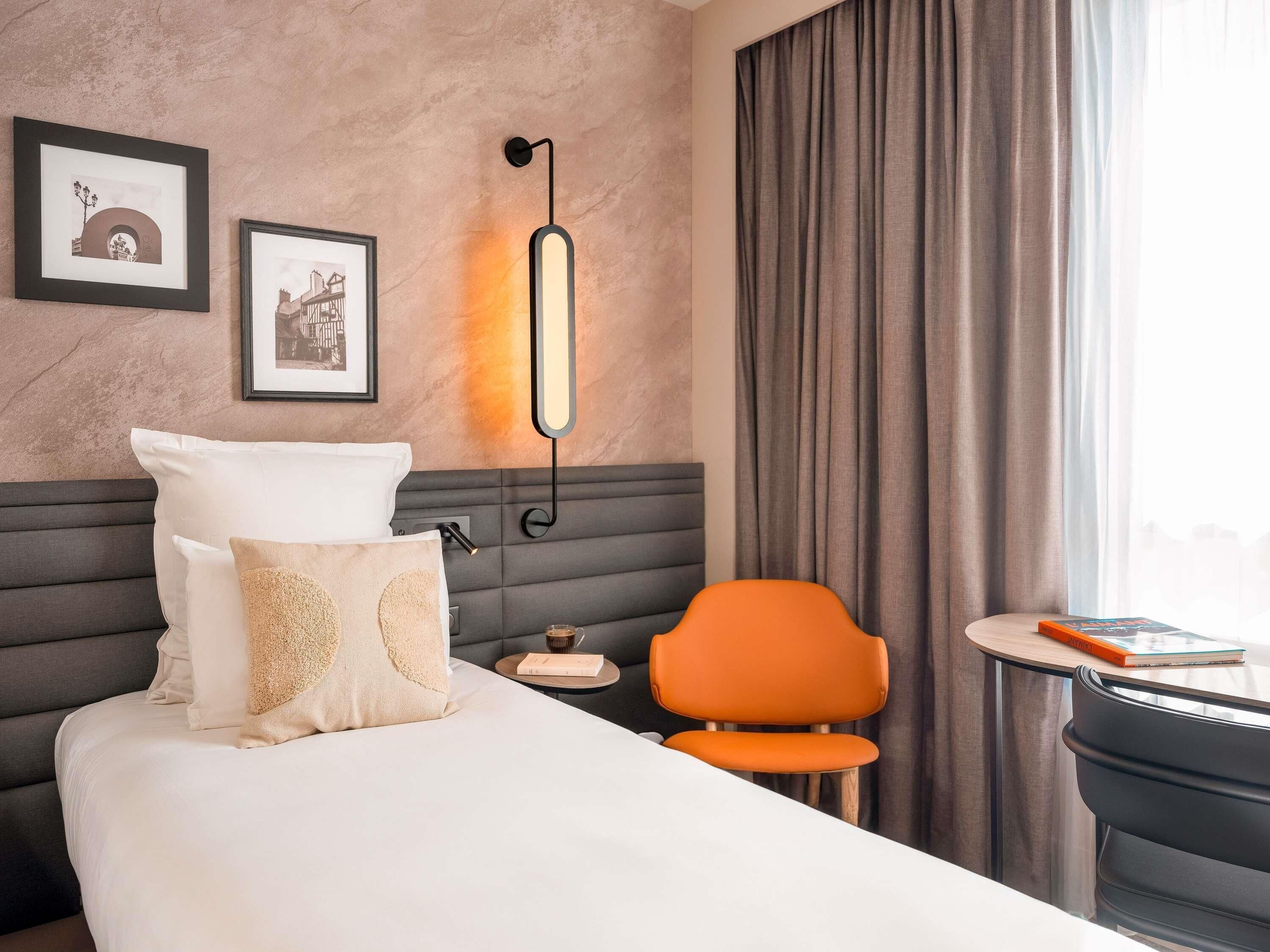 Una camera d'albergo con un letto, un comodino, una poltrona arancione, una lampada a parete e due quadri incorniciati.