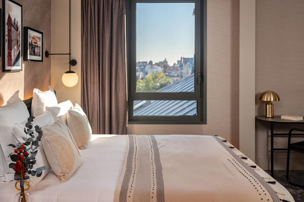 Ein Hotelzimmer mit einem großen Bett, Blick auf die Stadt durch ein Fenster, einer Lampe und einem kleinen Tisch ebenfalls mit einer Lampe.