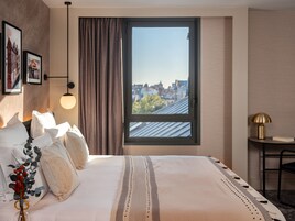 Una habitación de hotel con una cama grande, vistas de la ciudad a través de una ventana, una lámpara y una mesita con lámpara.