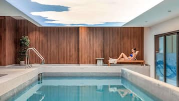 A modern indoor pool with a clear blue water surface, a wooden wall, and a person relaxing on a lounge chair.