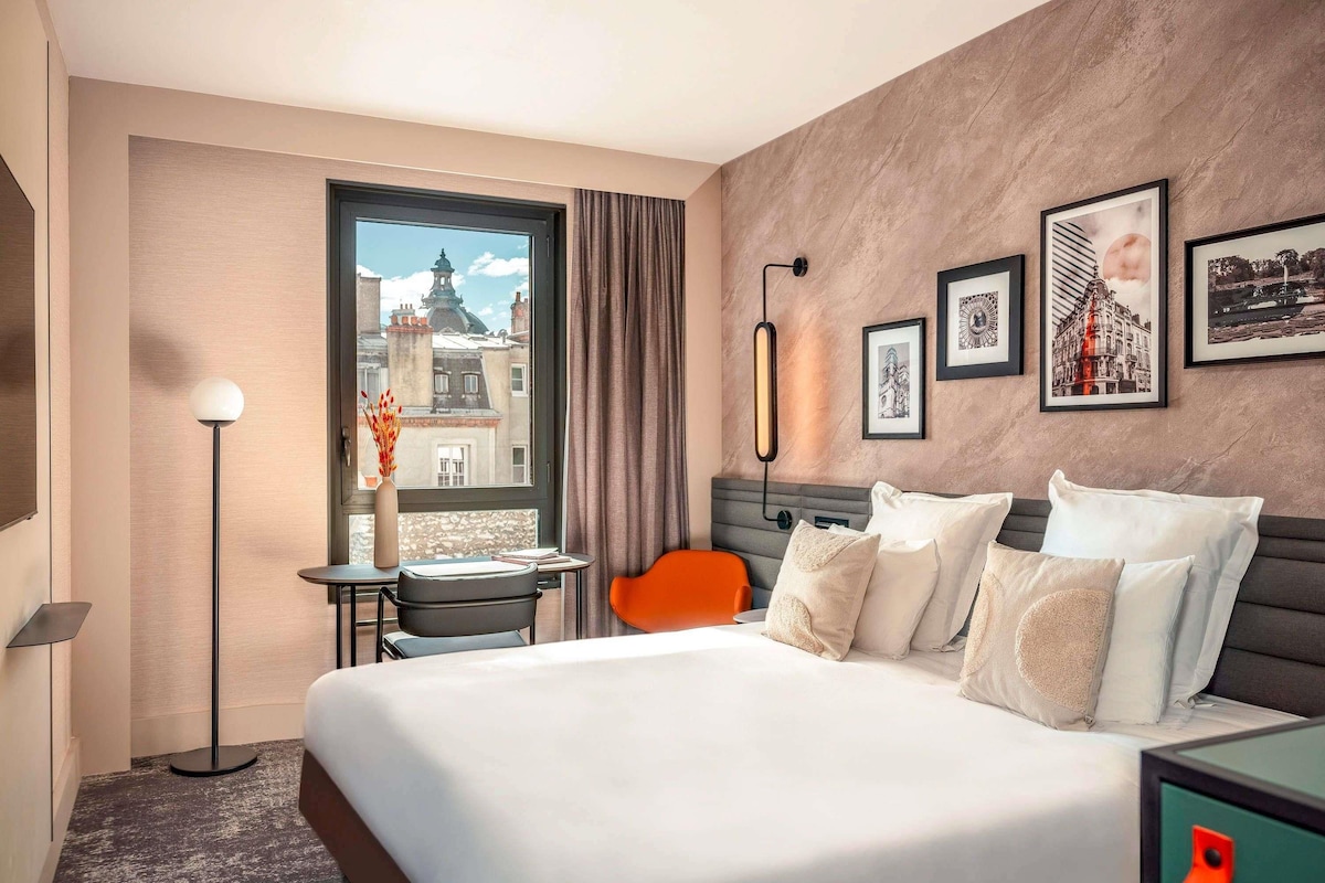 Habitación de hotel con una cama grande, un escritorio con una silla, una lámpara de pie, una ventana con vistas a edificios y cuadros en la pared.