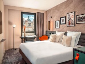 Une chambre d’hôtel avec un grand lit, un bureau avec une chaise, une lampe sur pied, une fenêtre donnant sur des immeubles et des tableaux encadrés accrochés au mur.