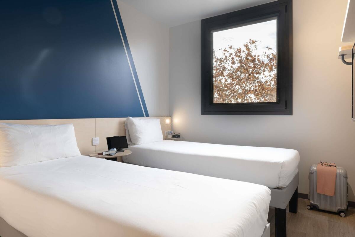 A hotel room with two beds, a desk with a computer, a window with a view of trees, and a suitcase on the floor.