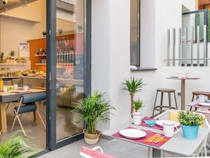 A modern dining area with a glass partition, a kitchen visible through the partition, and a small table with a plant and a mug.