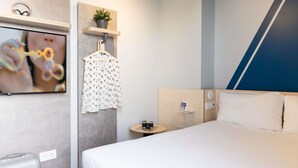 A modern hotel room with a large bed, a TV mounted on the wall, a small table with a camera, and a hanging garment. - Ibis Budget Valencia Centro Puerto (Valencia)