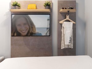 A modern room with a flat-screen TV mounted on a textured wall, a wooden shelf with a yellow bag and two potted plants, and a white towel hanging on a wooden hanger. - Ibis Budget Valencia Centro Puerto (Valencia)