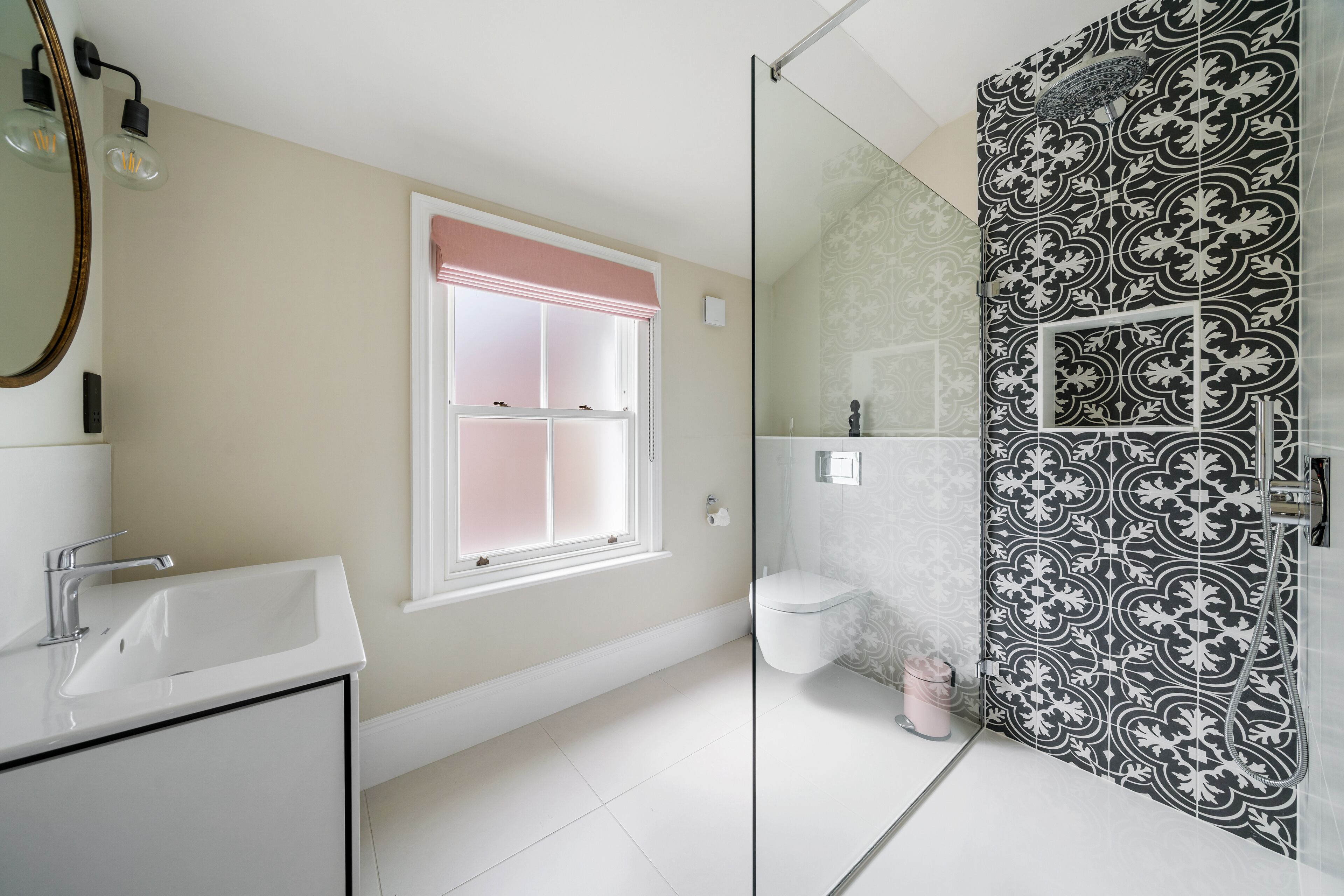 A bathroom with a white sink, a window with a pink blind, and a shower with black and white patterned tiles.