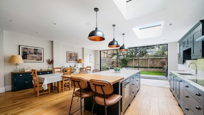 A modern kitchen with a large island, wooden dining table, and bar stools. There are pendant lights, a view to the backyard, and a skylight.