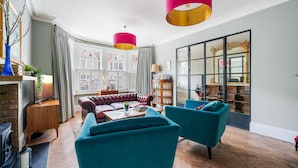 A living room with a fireplace, a red sofa, teal armchairs, and a large window with curtains.