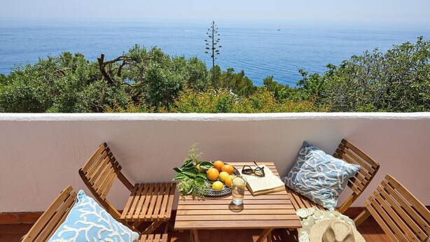 Une table en bois, sur laquelle se trouvent un panier de fruits, des verres et un livre, est placée sur un balcon donnant sur la mer.