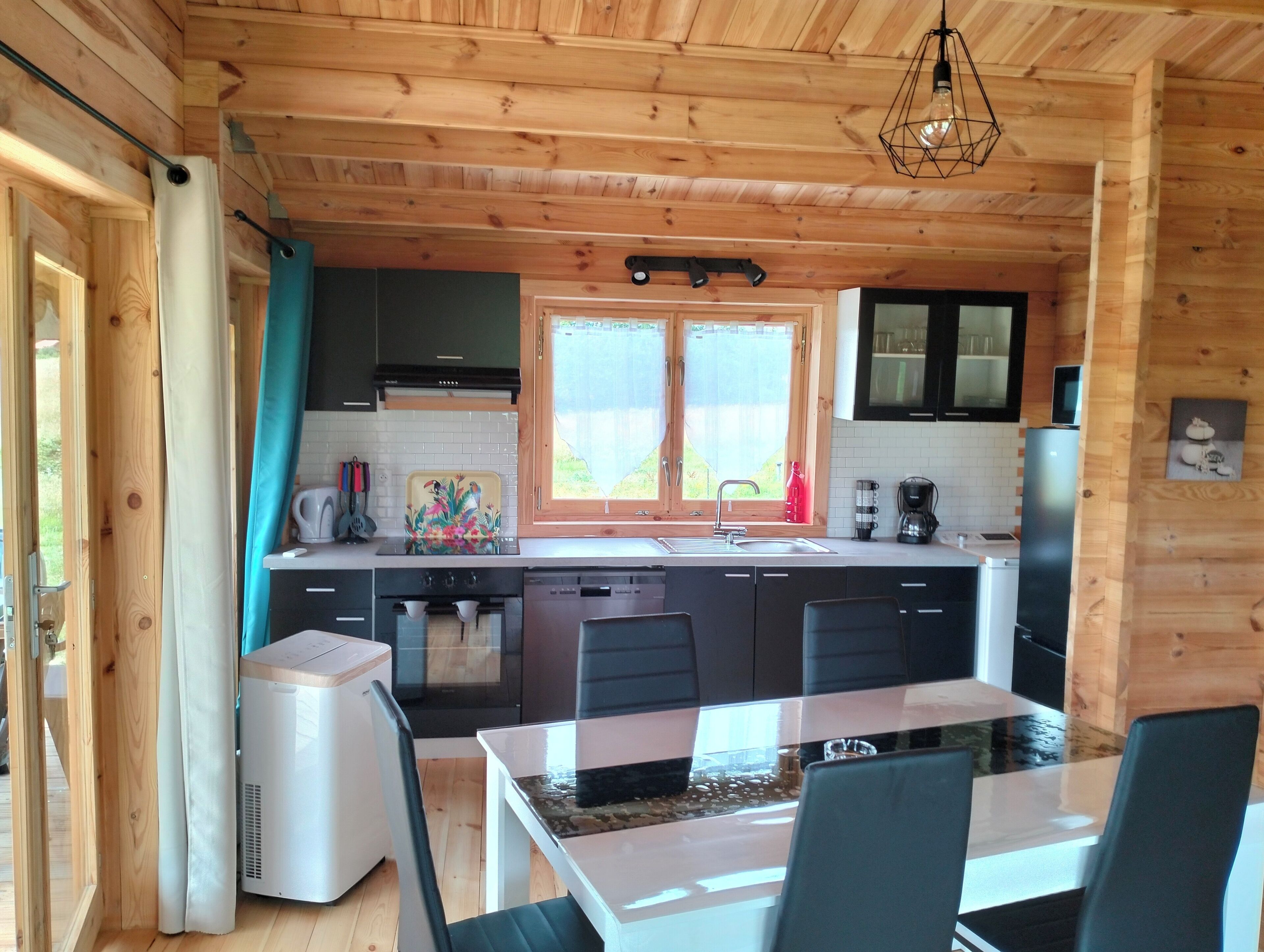 A wooden cabin-style kitchen with a dining table and chairs, a built-in oven, and a window with curtains.
