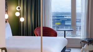 A hotel room with a bed, a red chair, a small round table, and a view of an airport. - Ibis Styles Rotterdam Ahoy (Rotterdam)