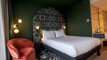 A hotel room with a large bed, a red chair, a patterned headboard, and a bedside table with a lamp.