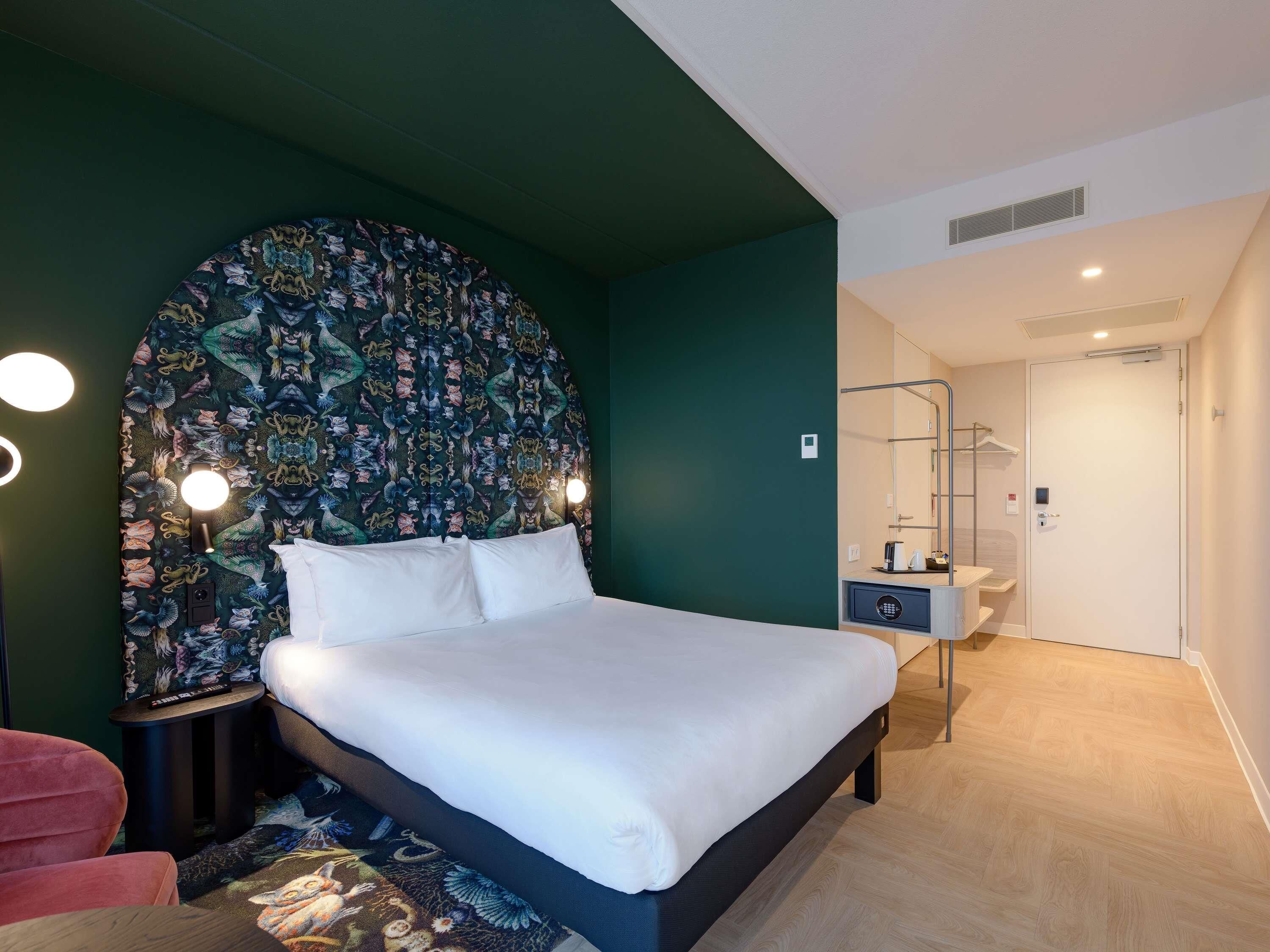 A modern hotel room with a large bed, a dark green accent wall, a patterned tapestry, and a small bedside table with a lamp.